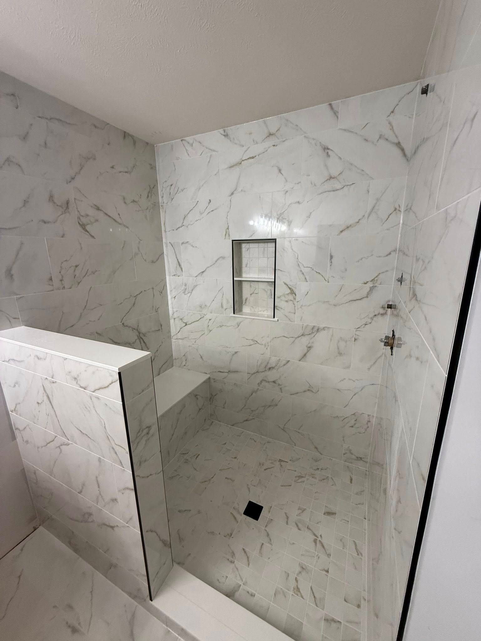 White marble-tiled shower with a built-in bench, shelf, and glass panel. Black drain and fixtures.