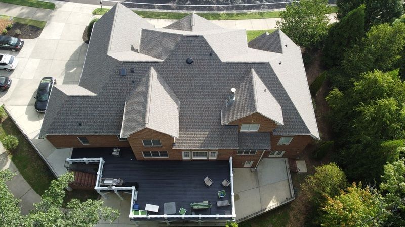 Overhead view of a large brick house with a complex gray roof, a dark deck, and surrounding greenery.