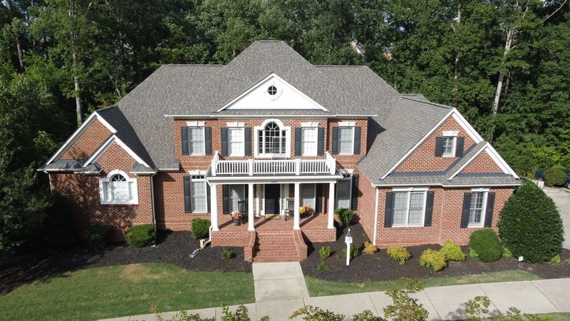 Two-story brick house with gray roof, black shutters, and front porch, surrounded by trees.