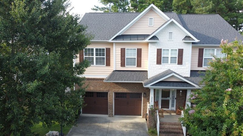 Two-story house with brick and tan siding, brown shutters and garage doors. Lush greenery surrounds.
