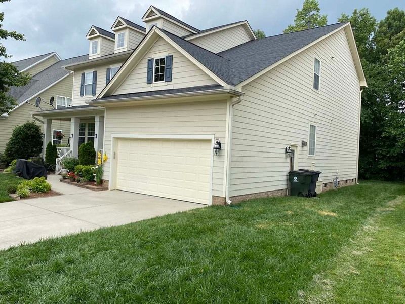 Two-story beige house with a garage, black roof, and green lawn. A trash can sits beside the house.