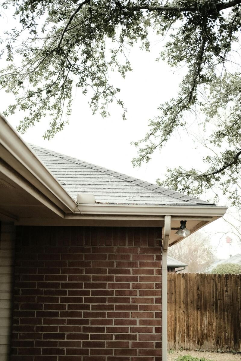Brick house corner with a roof, tan gutters, and tree branches against a cloudy sky.