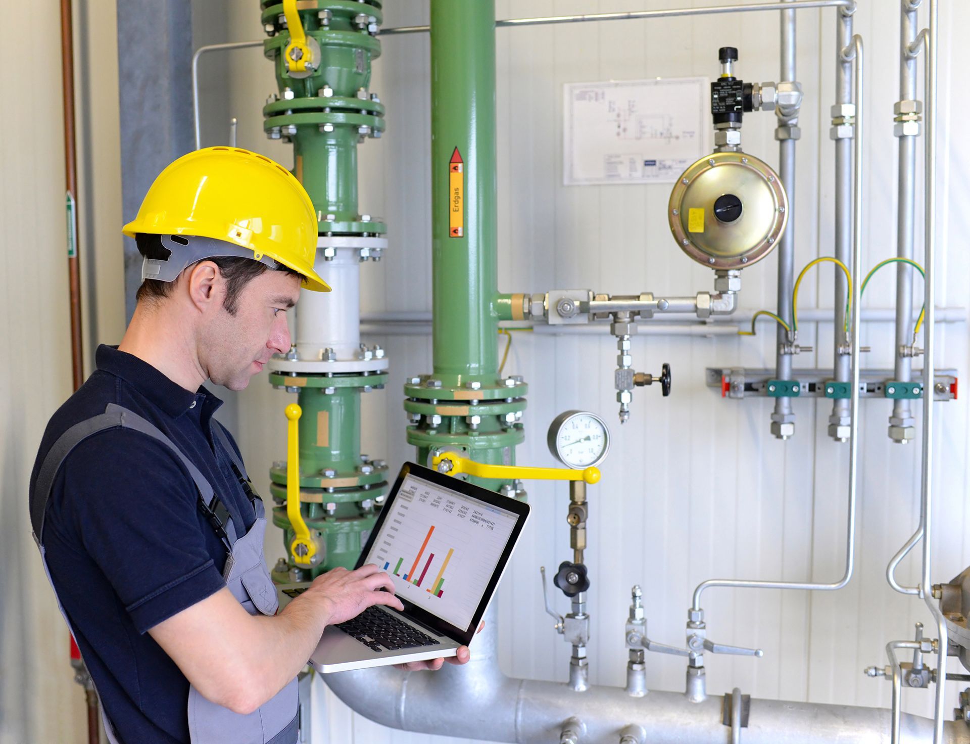 Un hombre que lleva un casco de seguridad está usando una computadora portátil frente a unas tuberías.