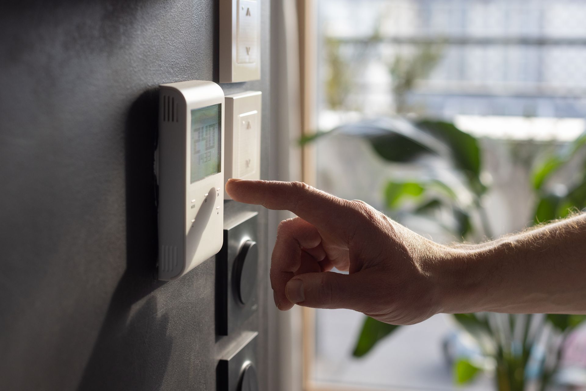Hand pressing a button on a white thermostat mounted on a dark wall, near a window with sunlight.