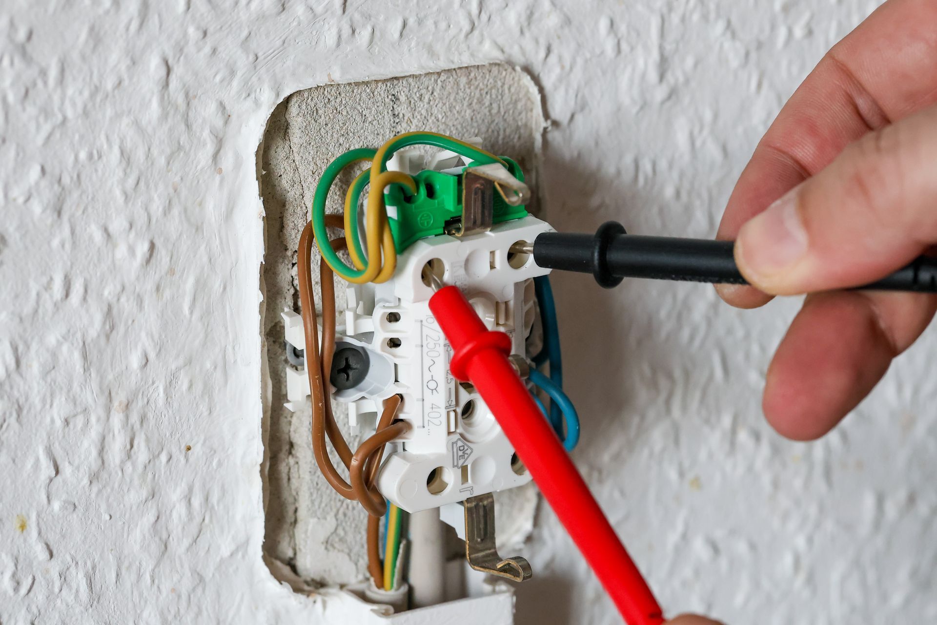 Hand testing an electrical outlet with red and black probes. Wires are visible, wall is white.