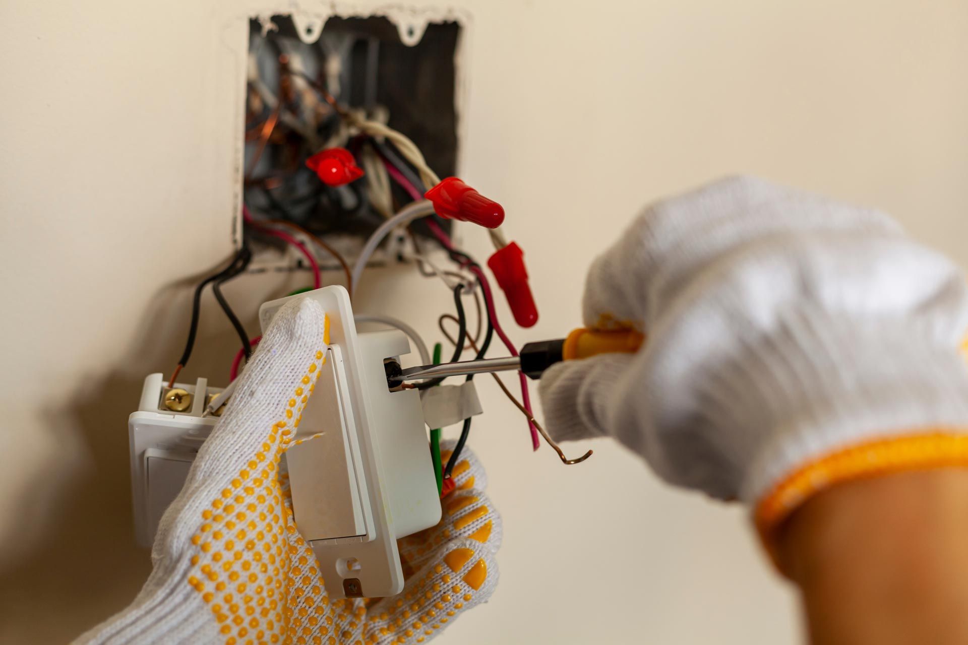 Gloved hands using a screwdriver to connect wires to a wall light switch.