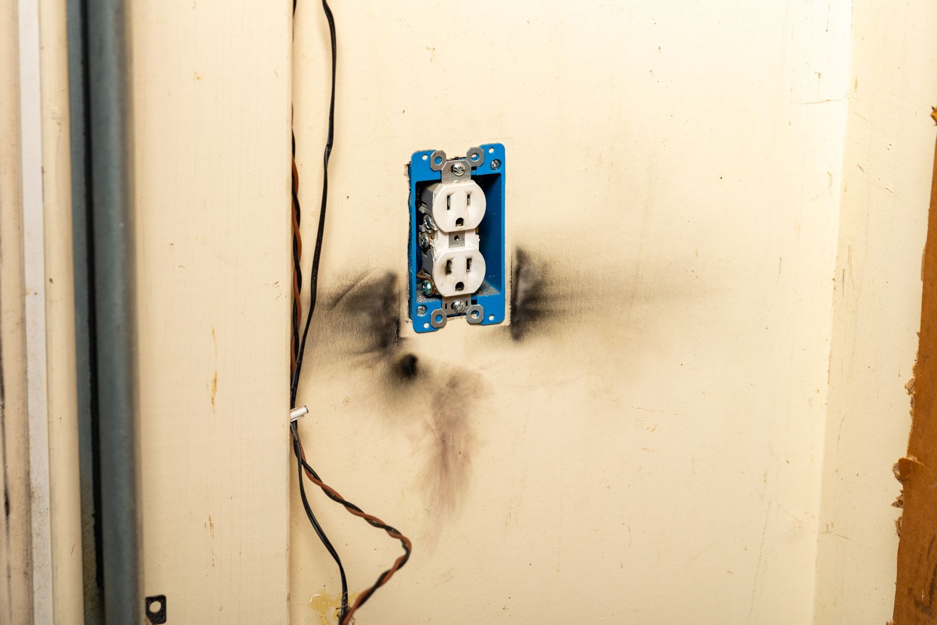 Electrical outlet with burn marks and exposed wiring on a beige wall.