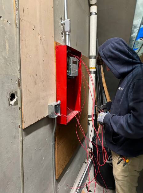 Hand testing an electrical outlet with red and black probes. Wires are visible, wall is white.