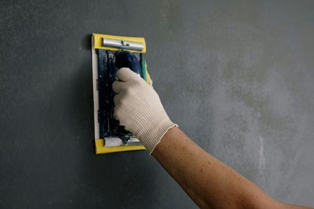 Person wearing a glove smoothing dark grey wall with a yellow-handled trowel.