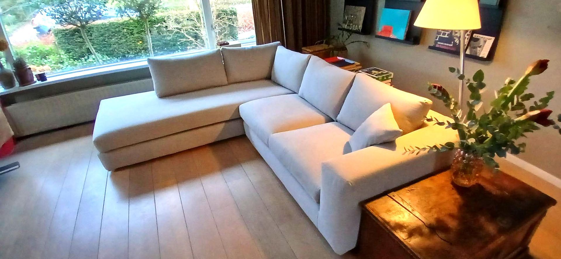 Een crèmekleurige hoekbank in een woonkamer met een houten salontafel en een groot raam.