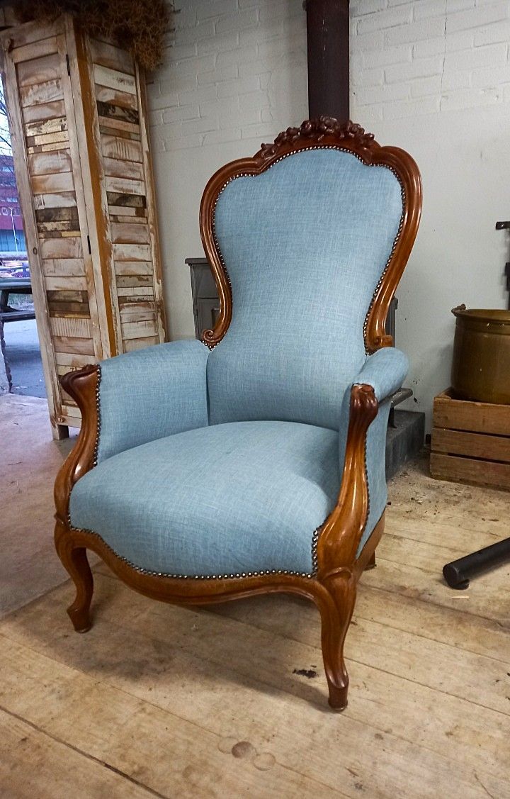 Lichtblauwe gestoffeerde fauteuil met een sierlijk houten frame, op een houten vloer, waarschijnlijk binnenshuis.