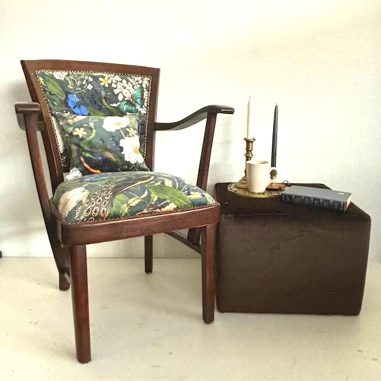 Een houten fauteuil met een stoffen bekleding met bloemenmotief en een kleine poef met kaarsen en een boek erop.