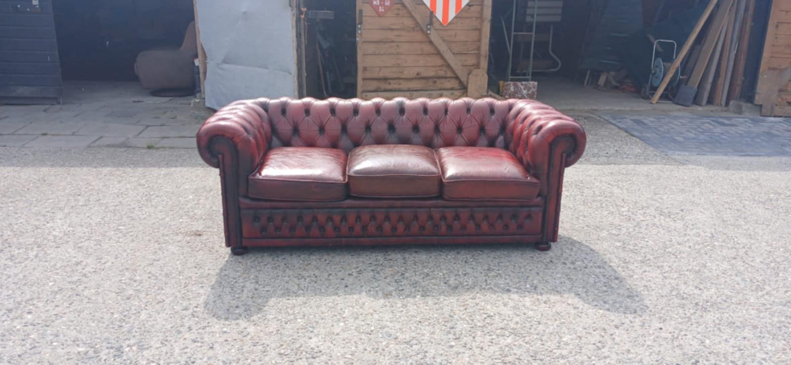 Rode leren Chesterfield-bank buiten op een grindoppervlak, voor een houten schuur.