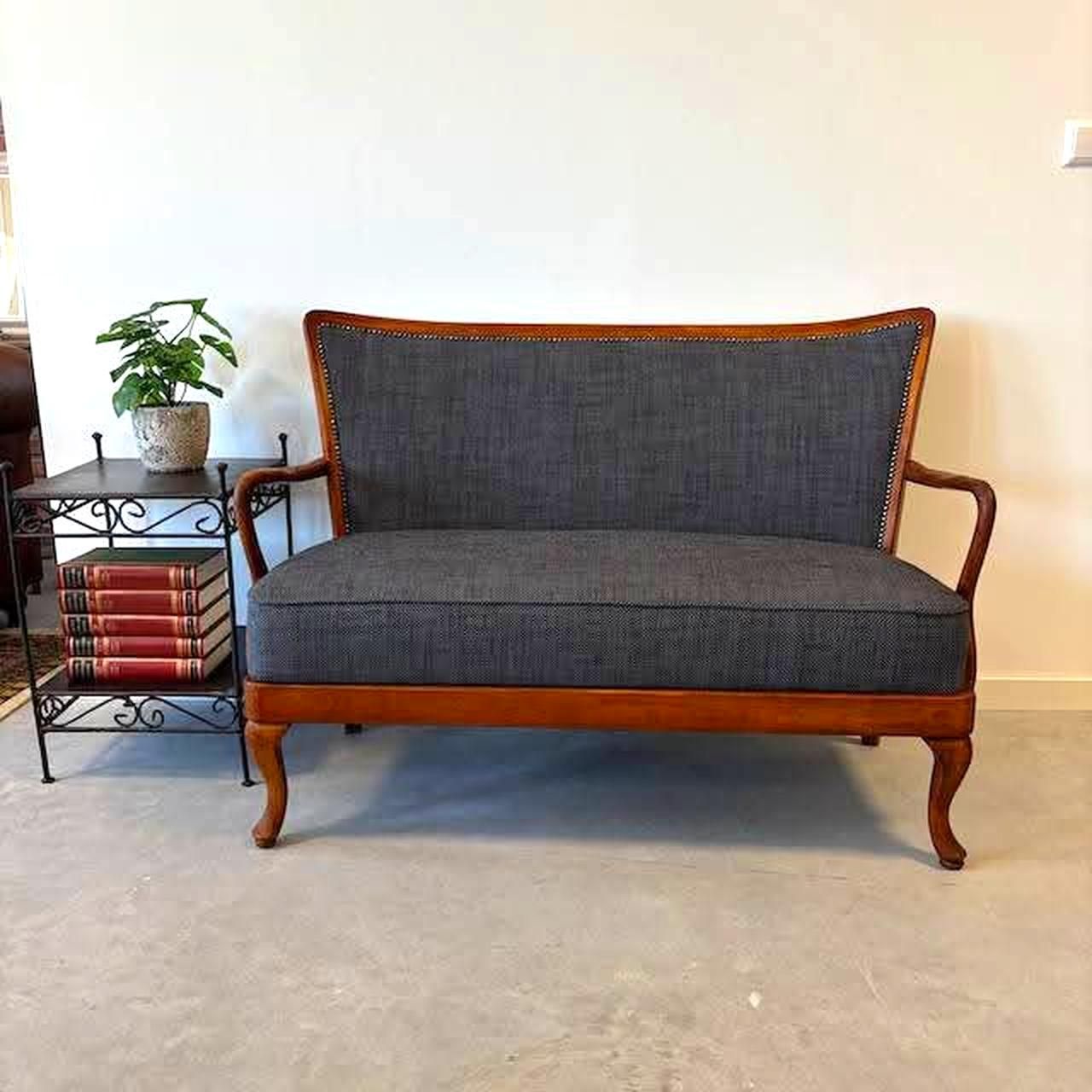 Antieke tweezitsbank met grijze bekleding en houten frame, naast een zwart bijzettafeltje met een plant en boeken.
