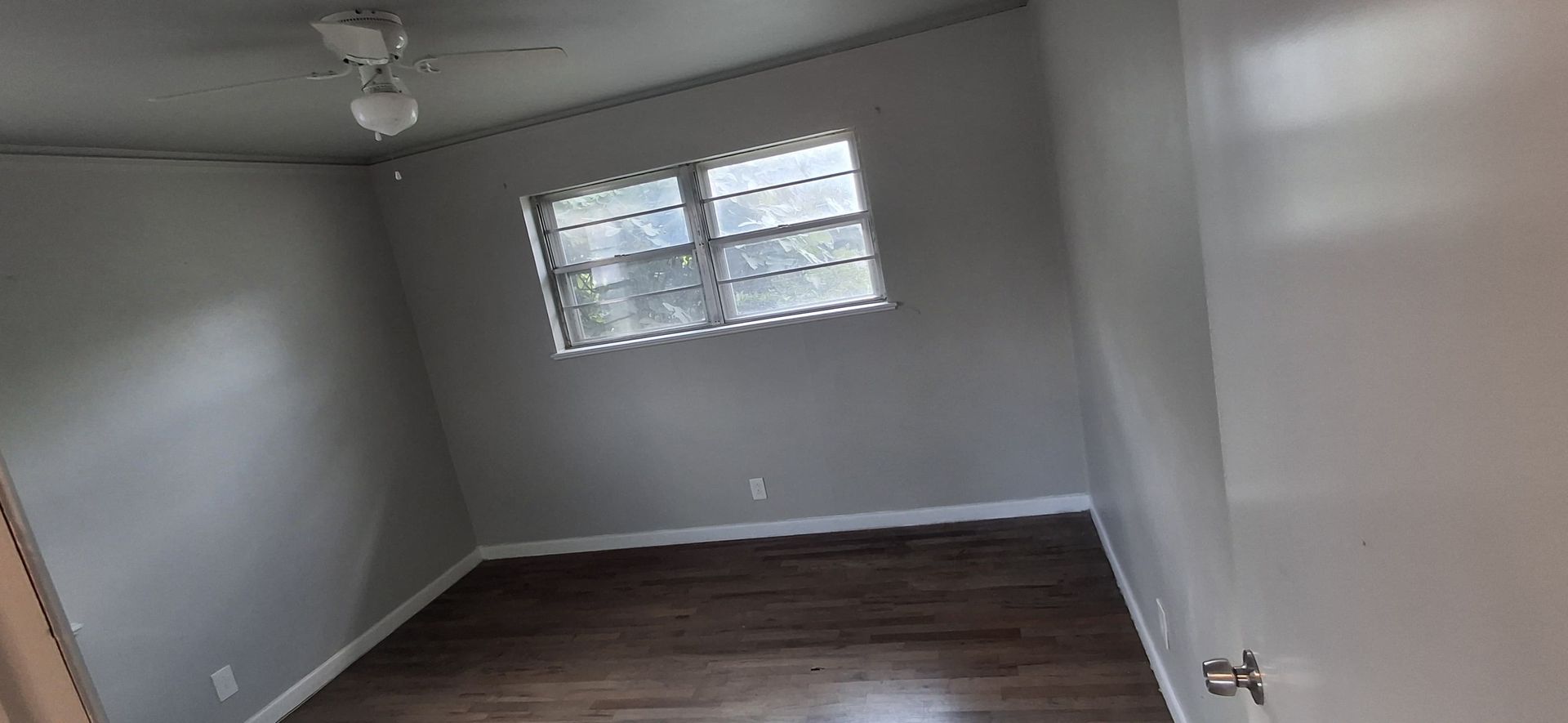 Empty bedroom with hardwood floors, a window, and a ceiling fan. Walls are painted gray.
