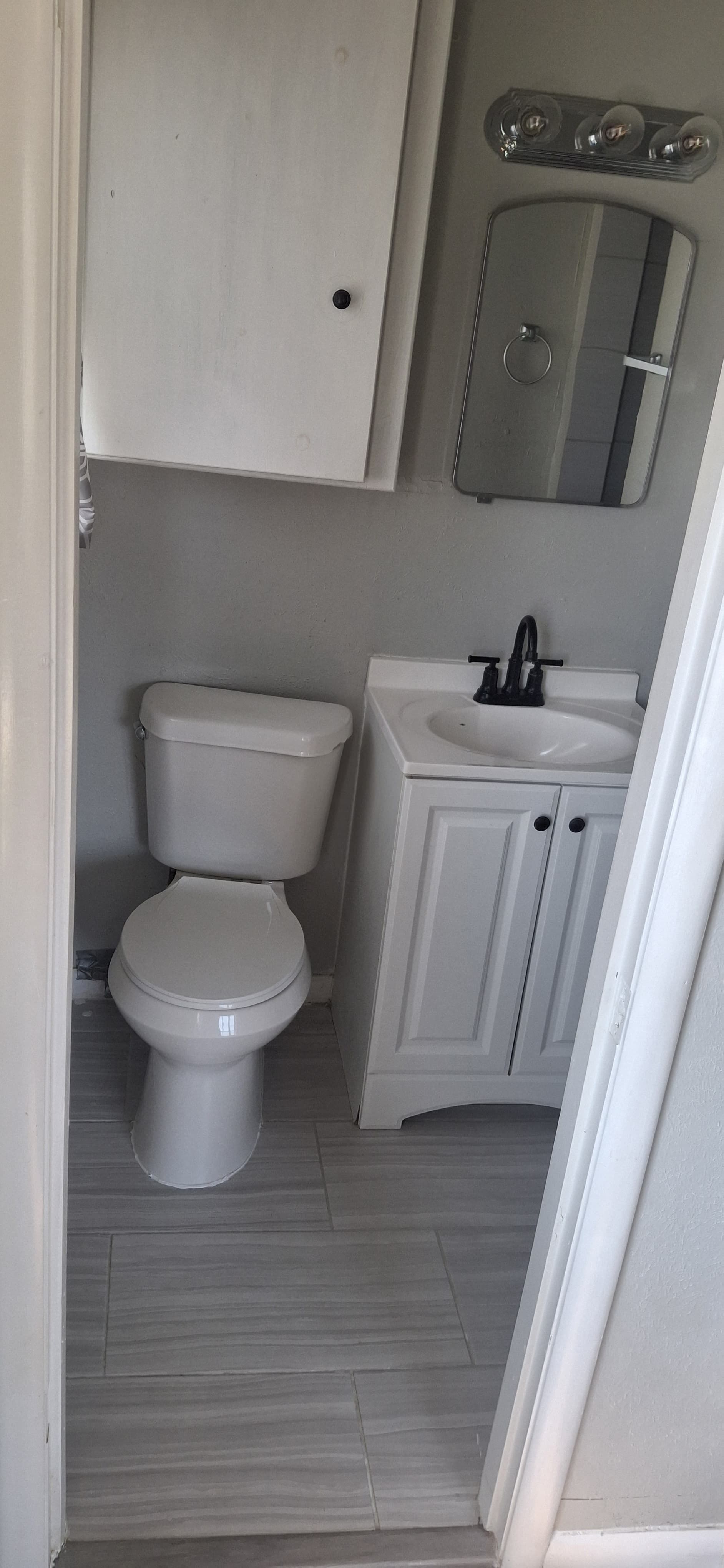 A small white bathroom with a toilet, sink, mirror, and cabinet. The floor is grey.