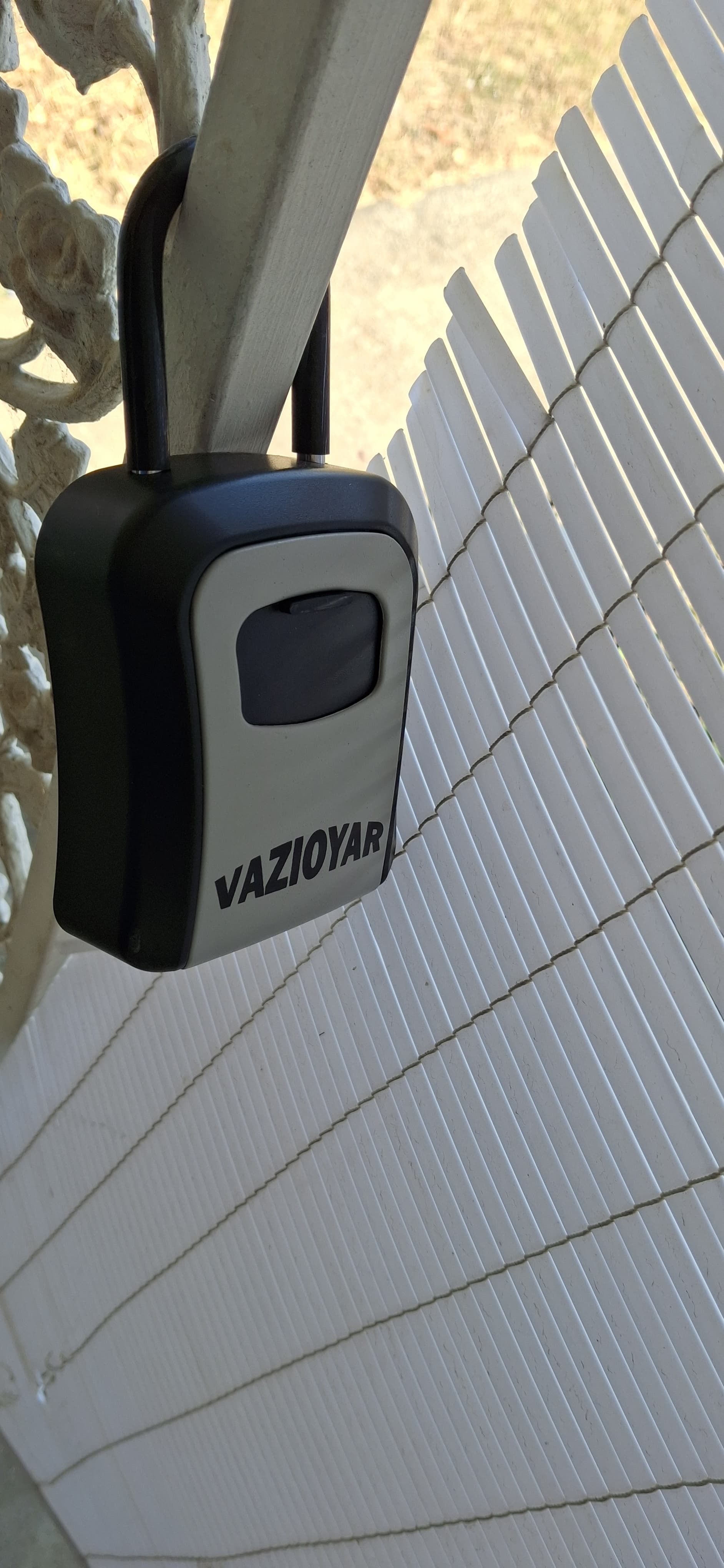 A black key safe hanging on a fence in the sunlight. The key safe is labeled 