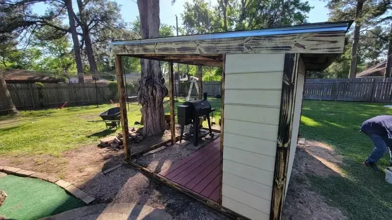 Backyard shelter with a grill and person working. Wooden structure with a red deck and white siding.
