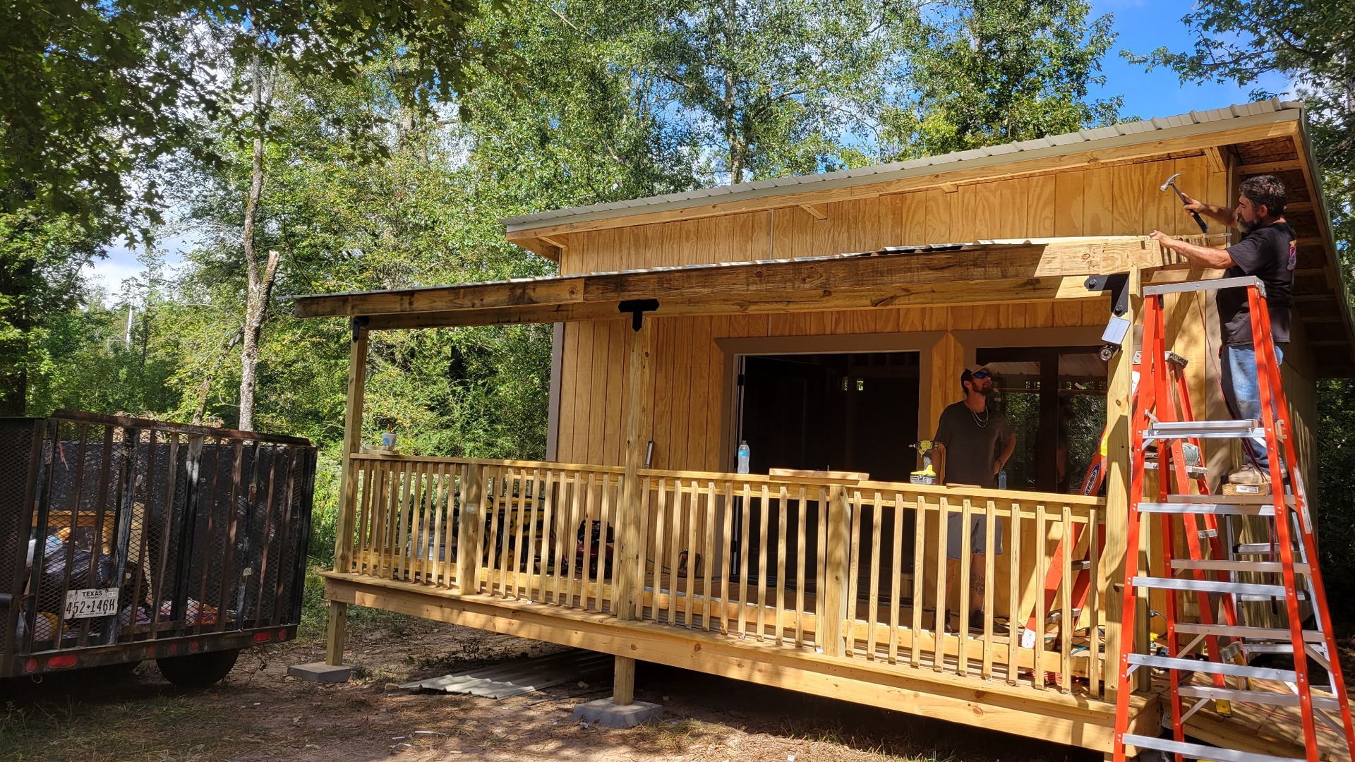 Construction of a wooden cabin with porch; two workers, one on a ladder, are building it outdoors.