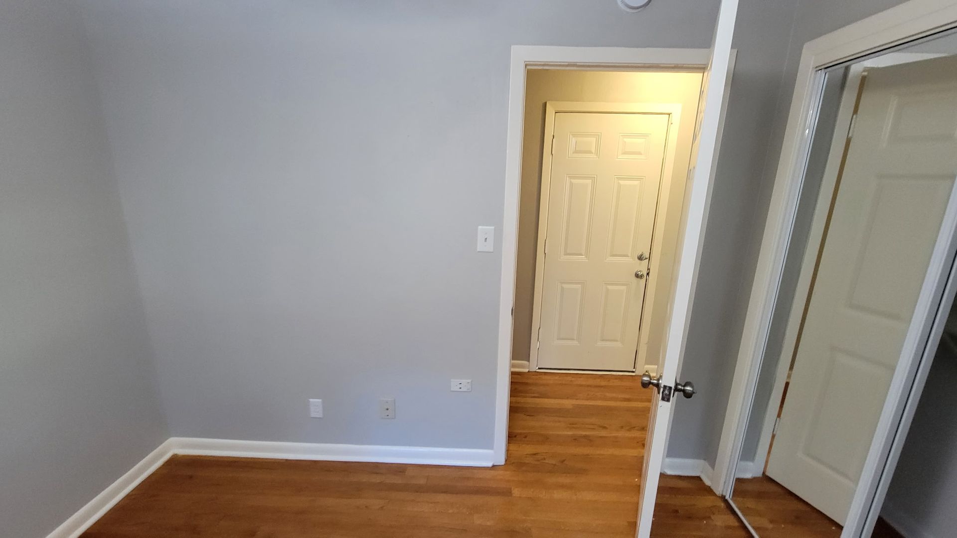 Empty room with wood floors and a doorway leading to another room. White door, light gray walls.