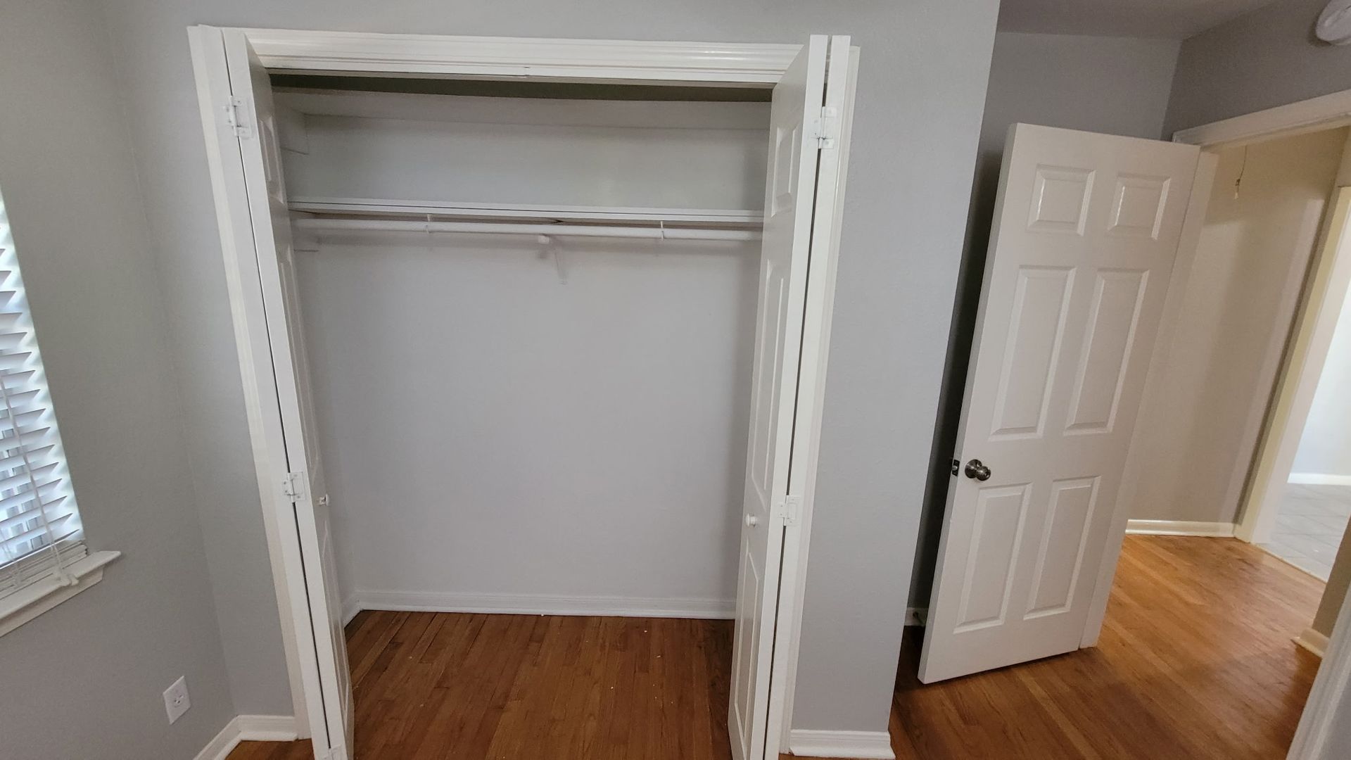 Empty white closet with open doors; hardwood floor, interior door visible.
