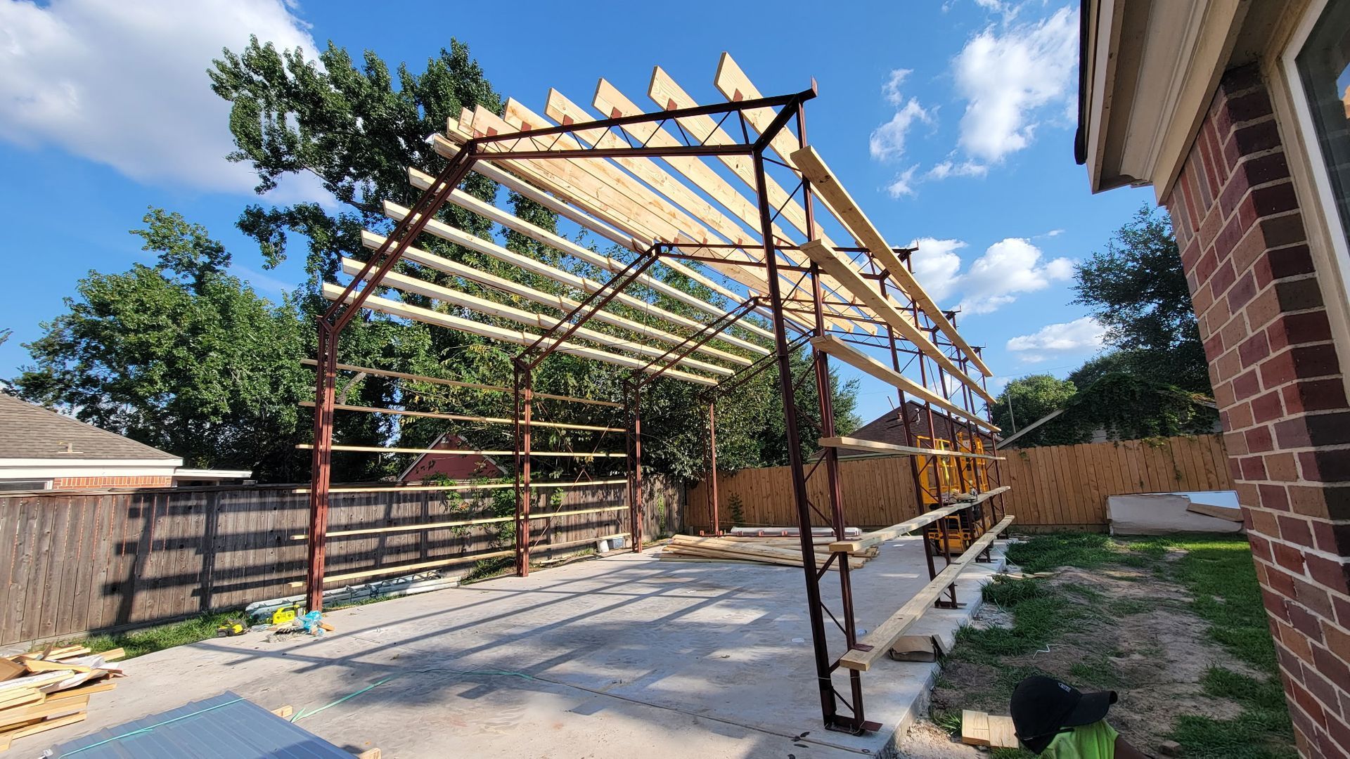 Steel frame carport under construction; wood beams laid across the top. Sunny day, backyard setting.