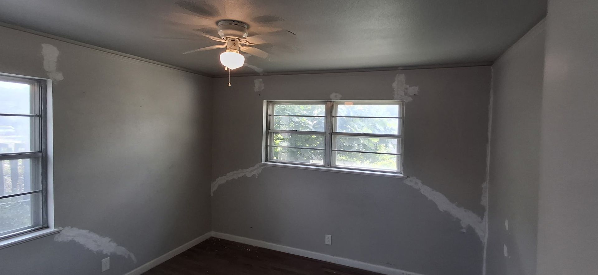 An empty room with gray walls and window. Drywall patching visible. Ceiling fan.