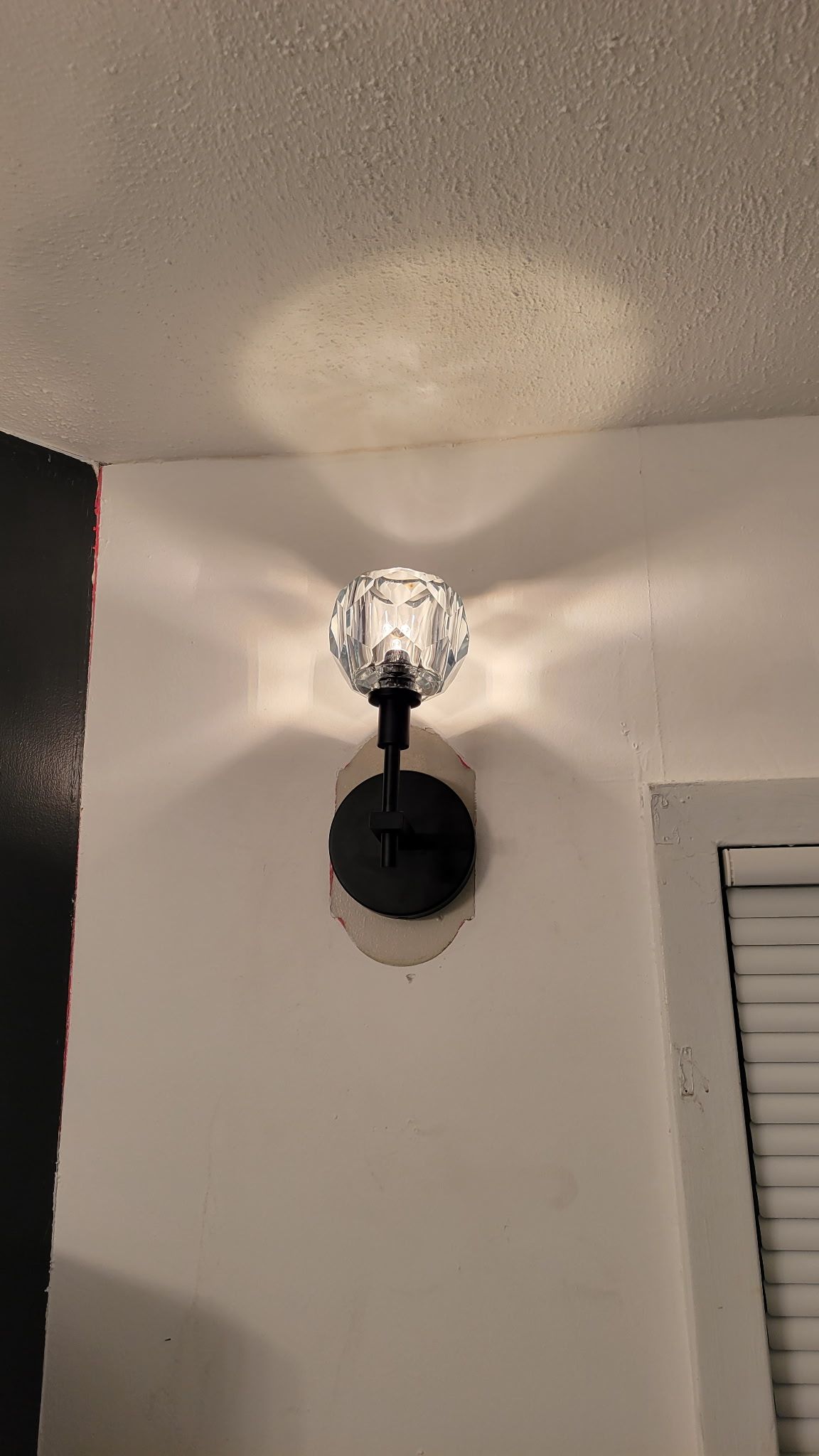 A crystal wall sconce emits light on a white wall. Black fixture attached.