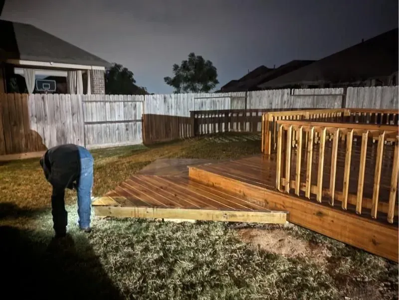 Person working on a wooden deck in a backyard at night. Deck is brown, fence is light-colored.