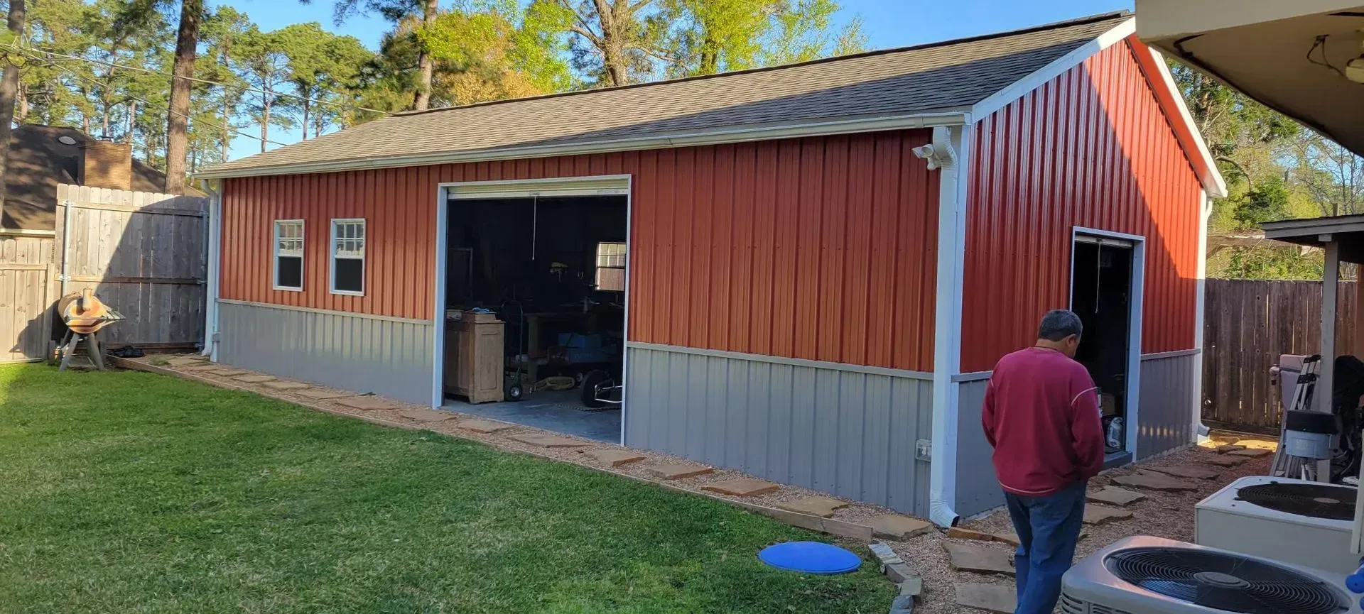 Red and gray metal shed with open garage door. Man in red shirt enters a doorway. Green grass.