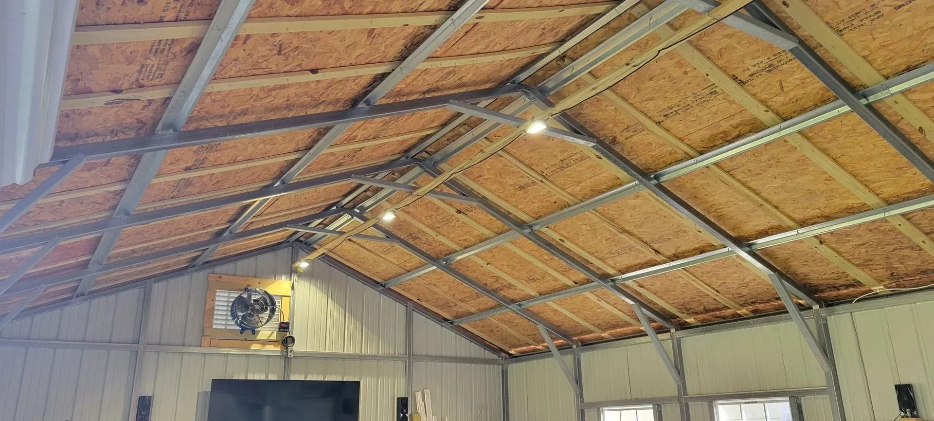 Interior view of a building's ceiling, with exposed beams and plywood panels. Three lights illuminate the space.