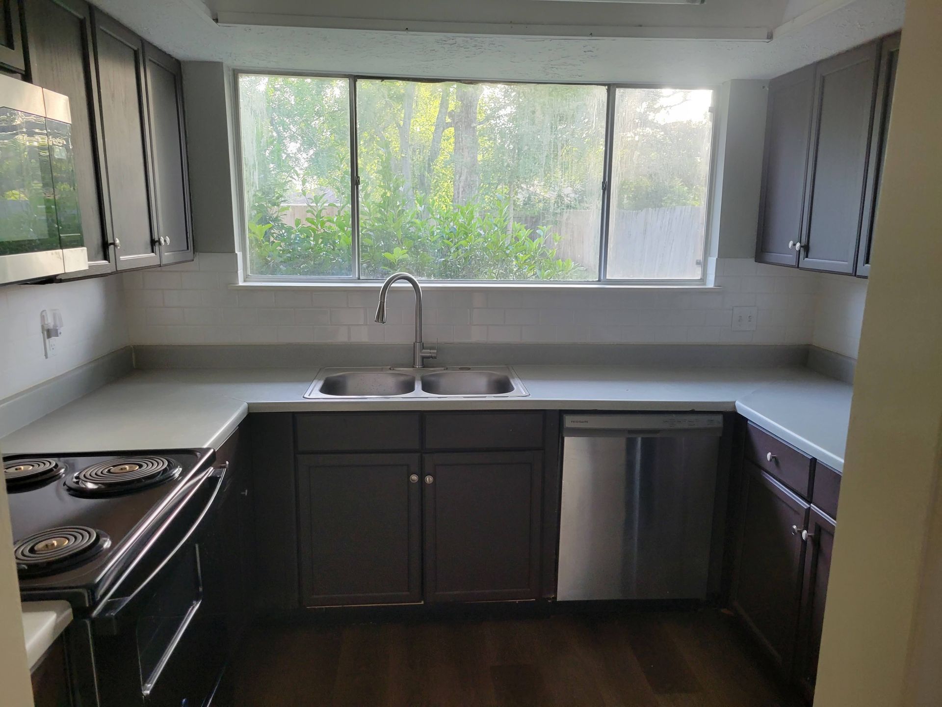 Dark gray kitchen cabinets with a stainless steel sink, and dishwasher. Bright window looking out onto greenery.