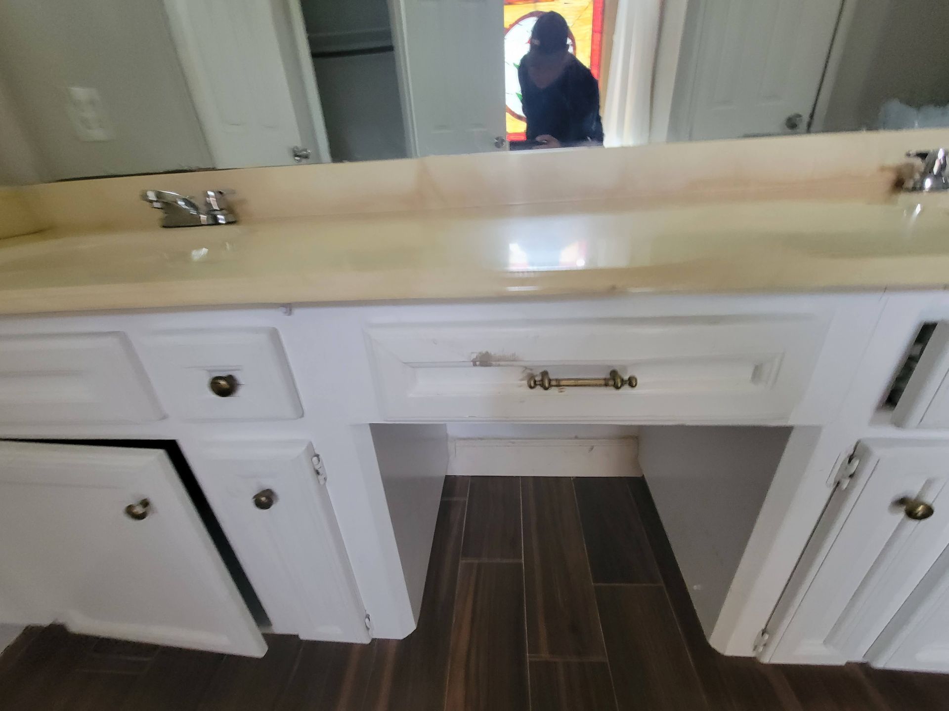 White bathroom vanity with cream-colored countertop and dark wood floor. A person is visible in the mirror.