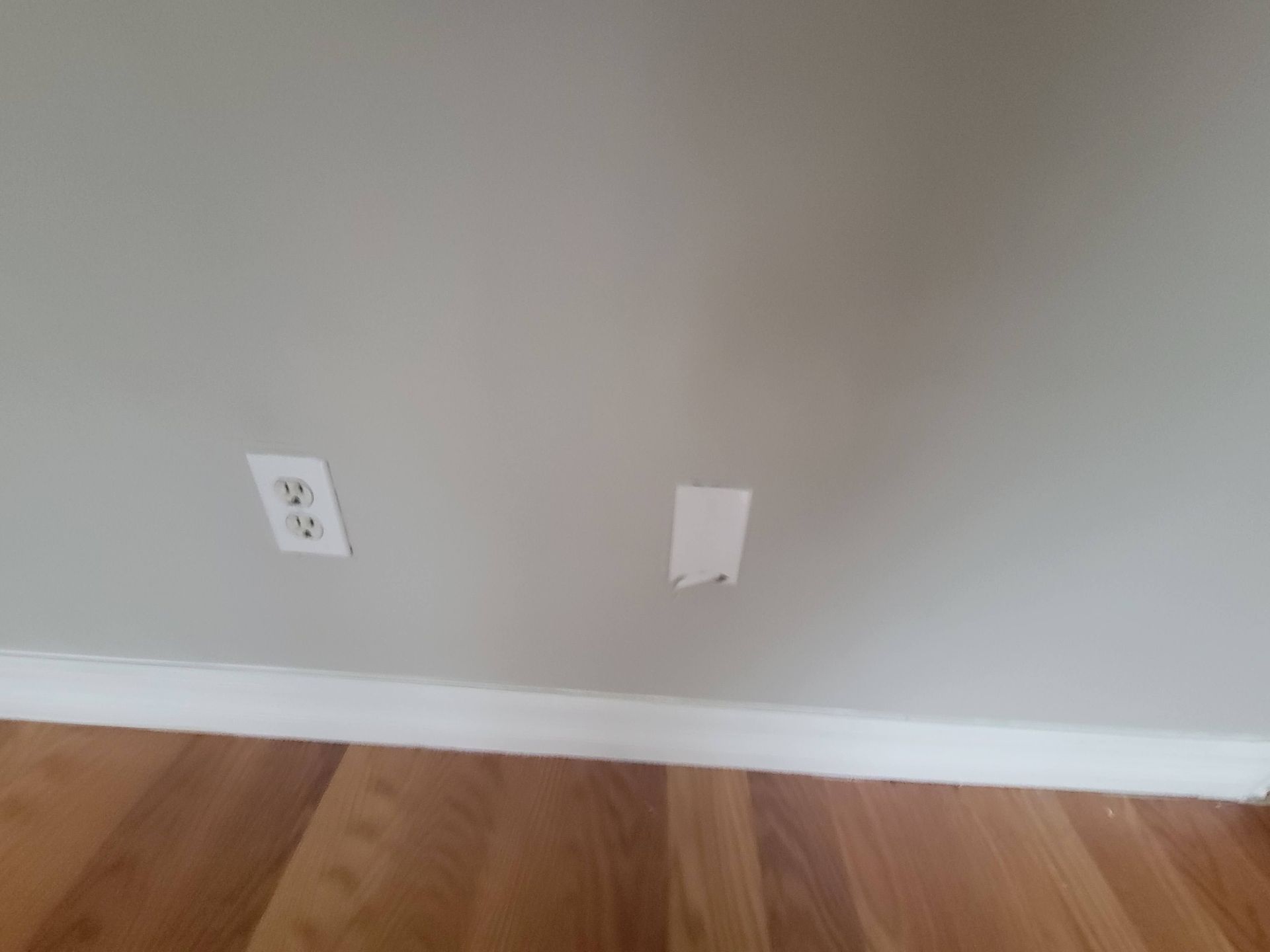 Gray wall with an electrical outlet and a rectangular hole, above a wood floor with white baseboard.