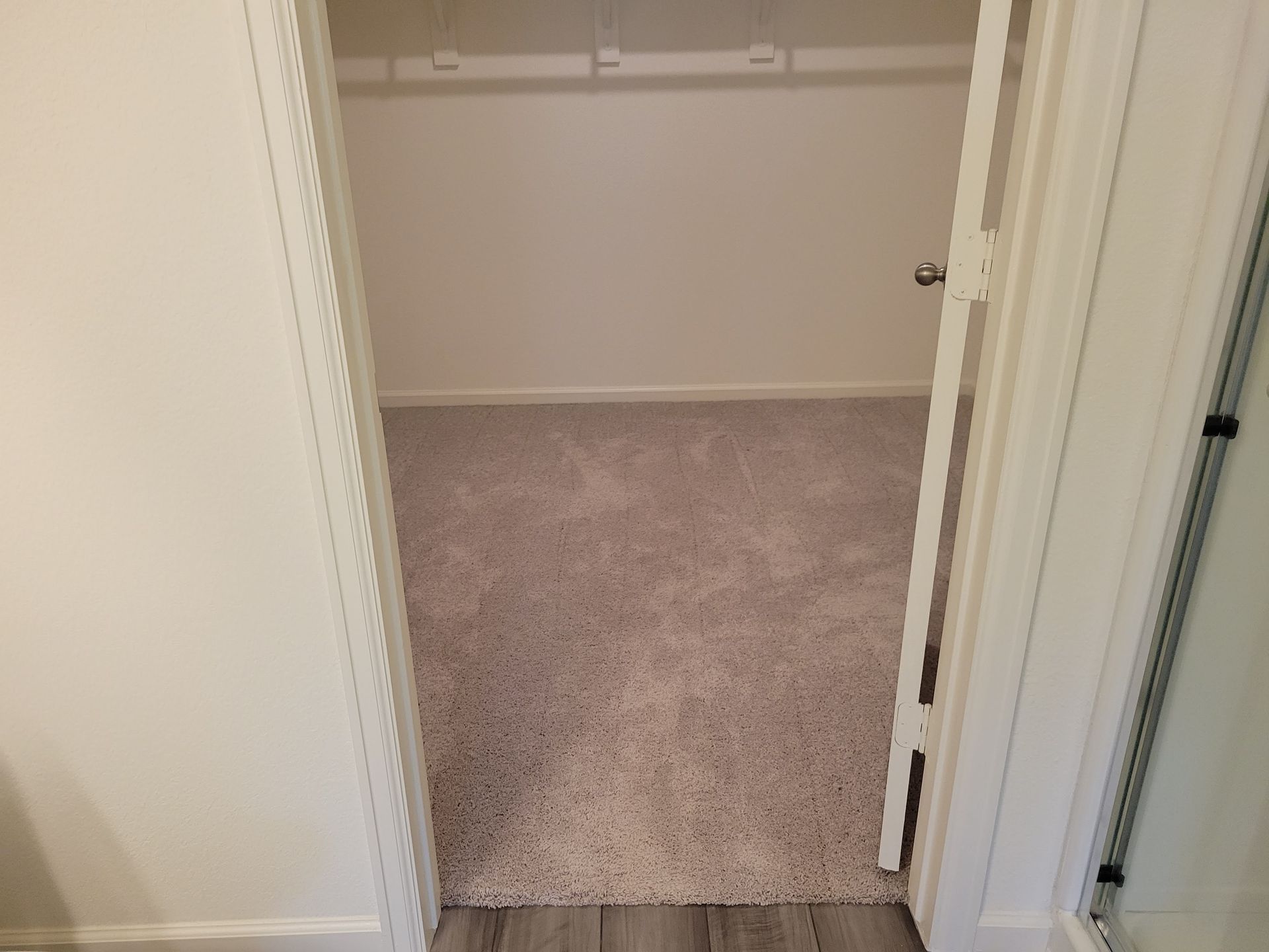 Empty walk-in closet with beige carpet and white walls, viewed through an open doorway.