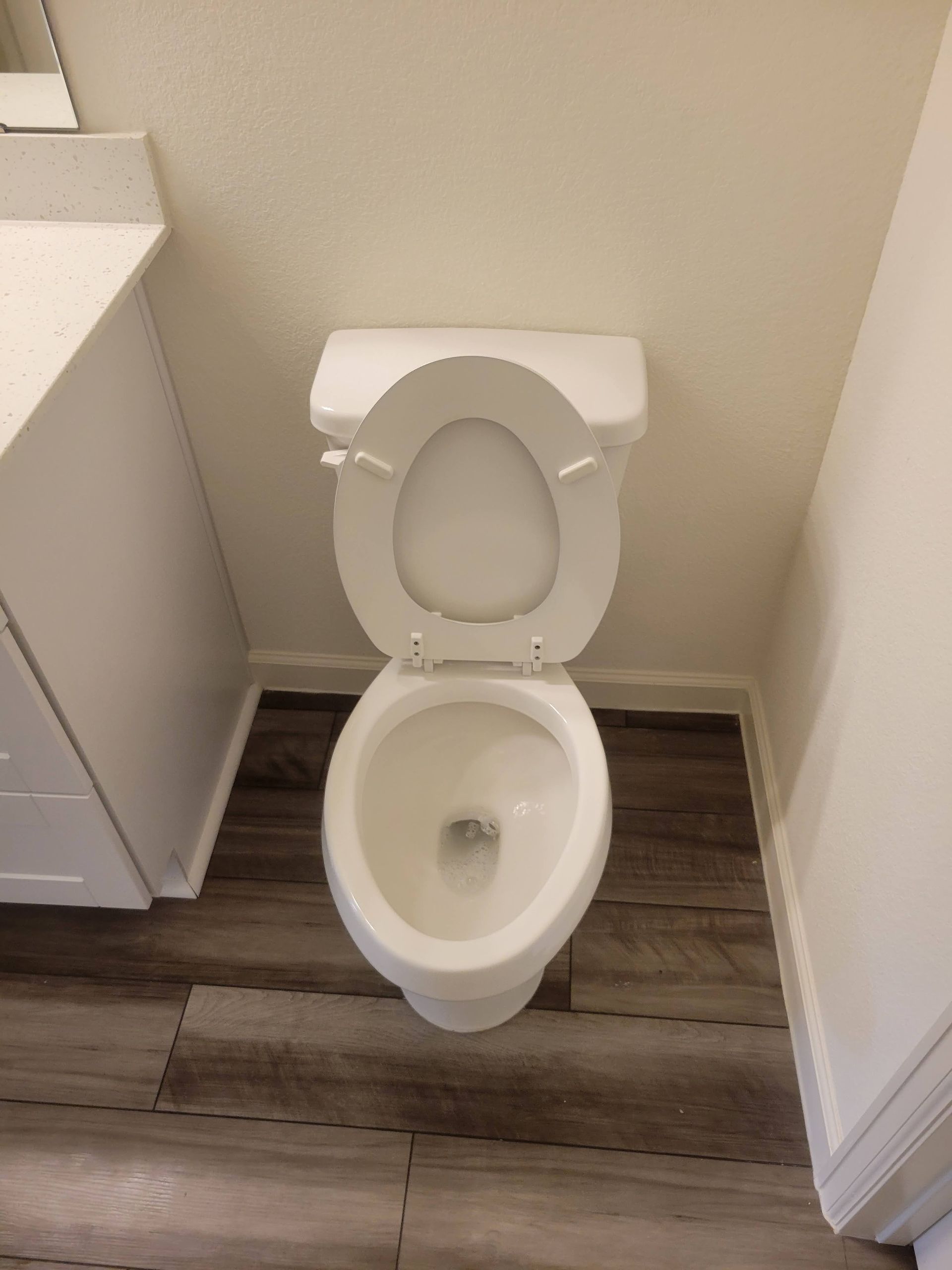 White toilet with open seat, on wood-look floor, against a white wall in a bathroom.