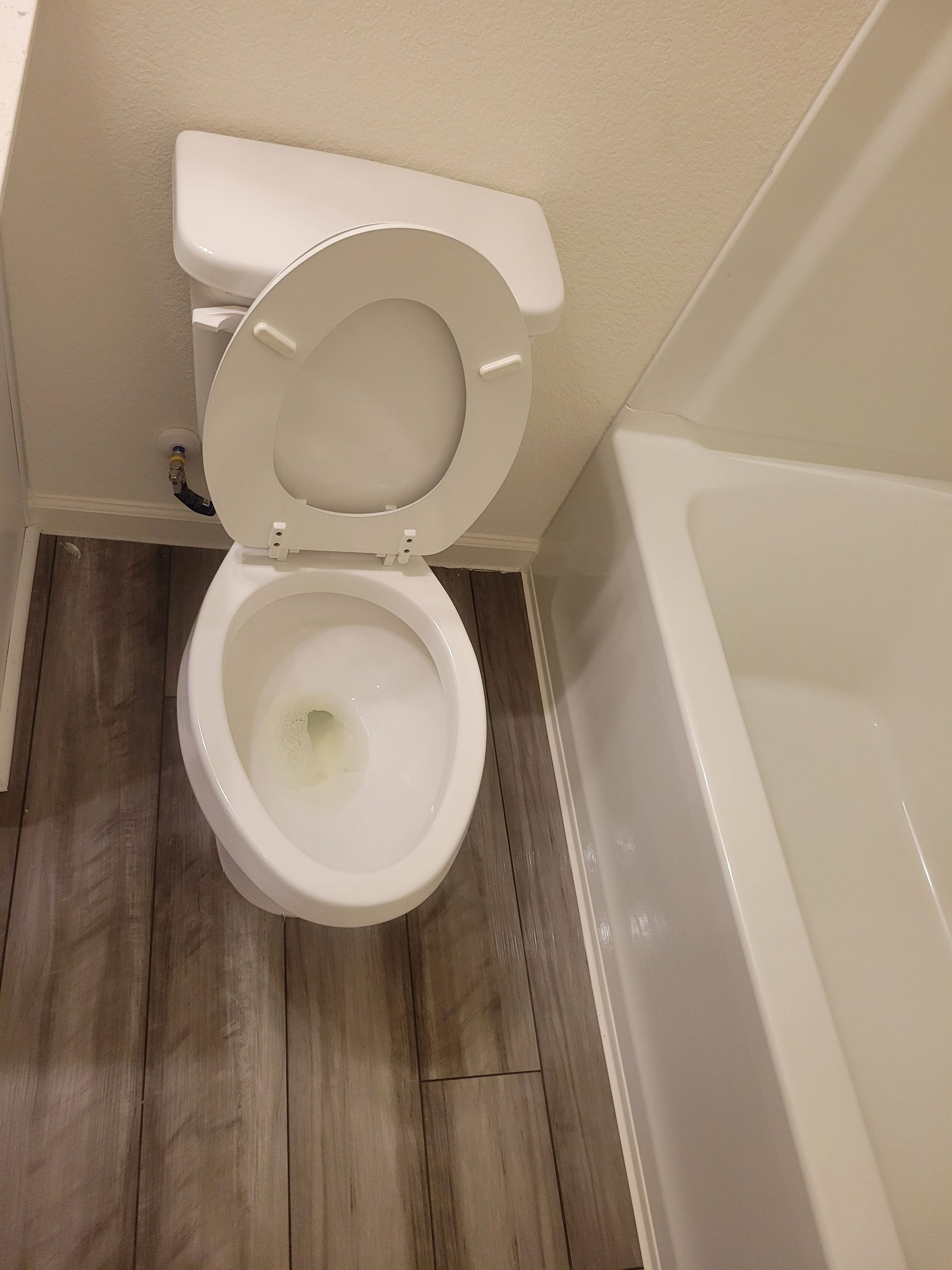 Toilet with the lid up, next to a white bathtub. The floor is wood-look gray tile.