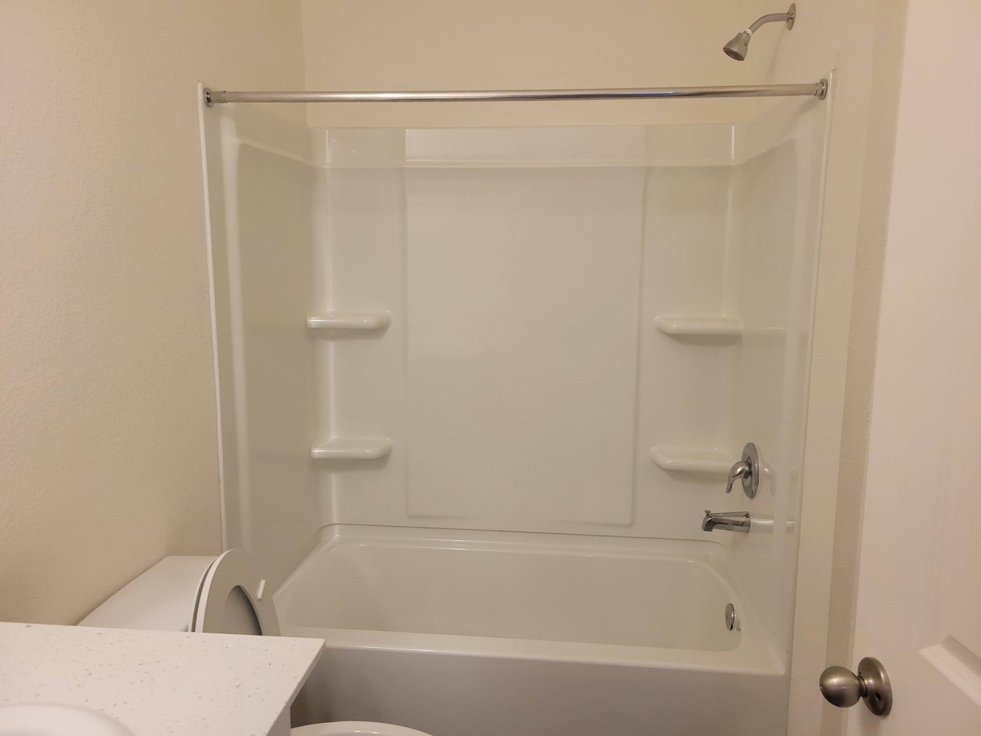 White bathroom with tub, showerhead, and two shelves; toilet and door in view.
