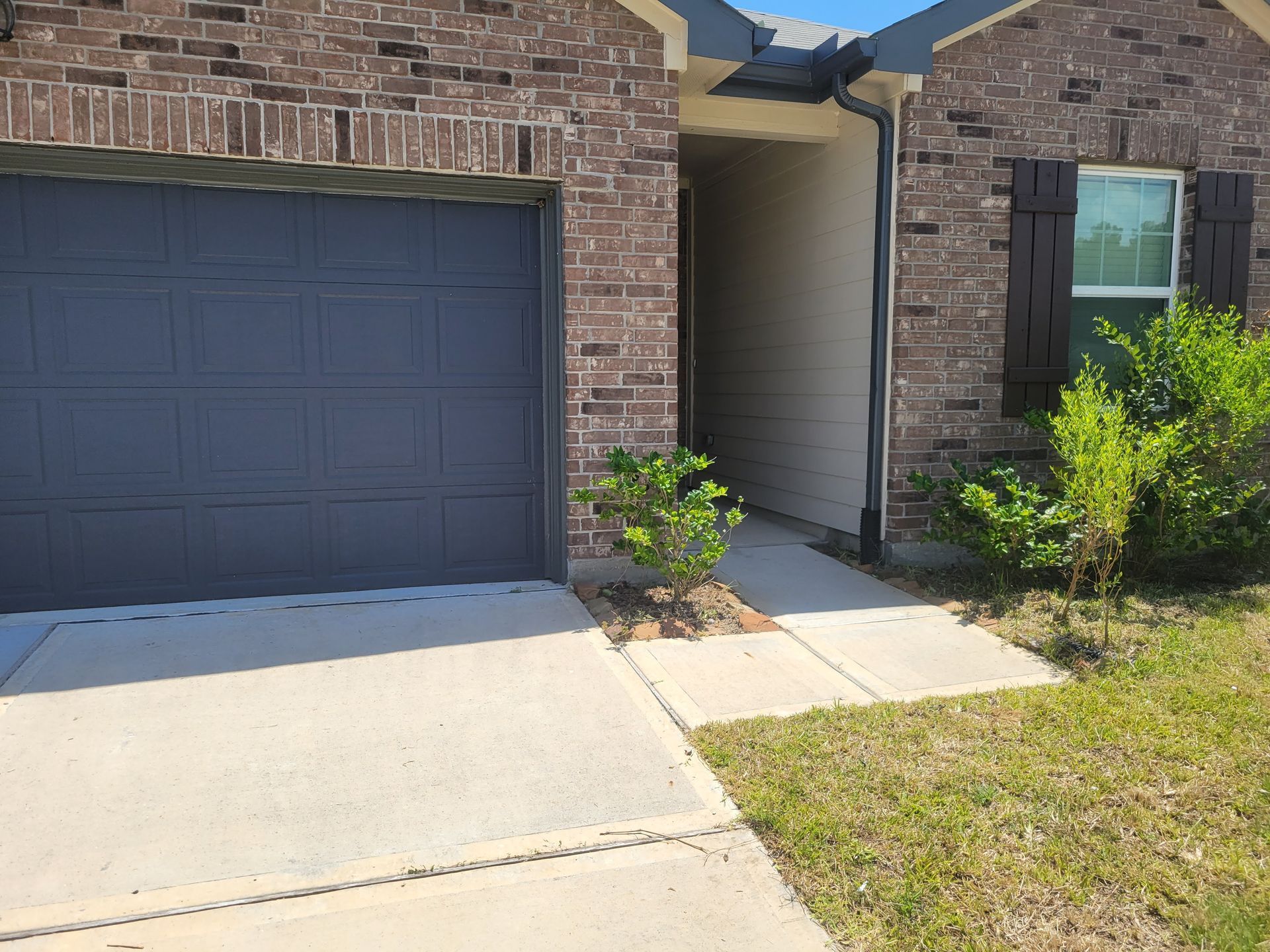 Brick house exterior with a gray garage door, walkway, and landscaping.