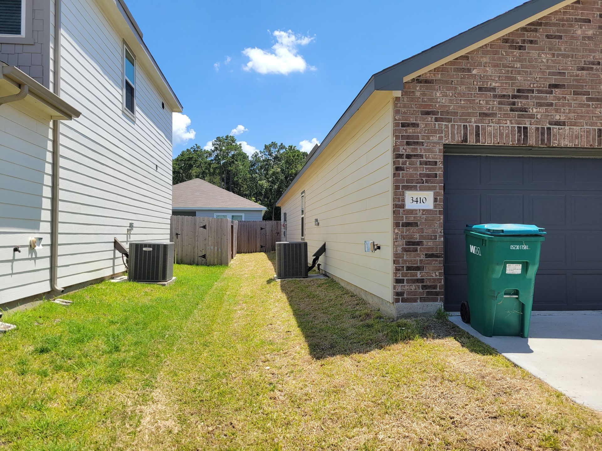 A narrow grassy side yard between two houses. The sky is blue. A trash can is by the garage.