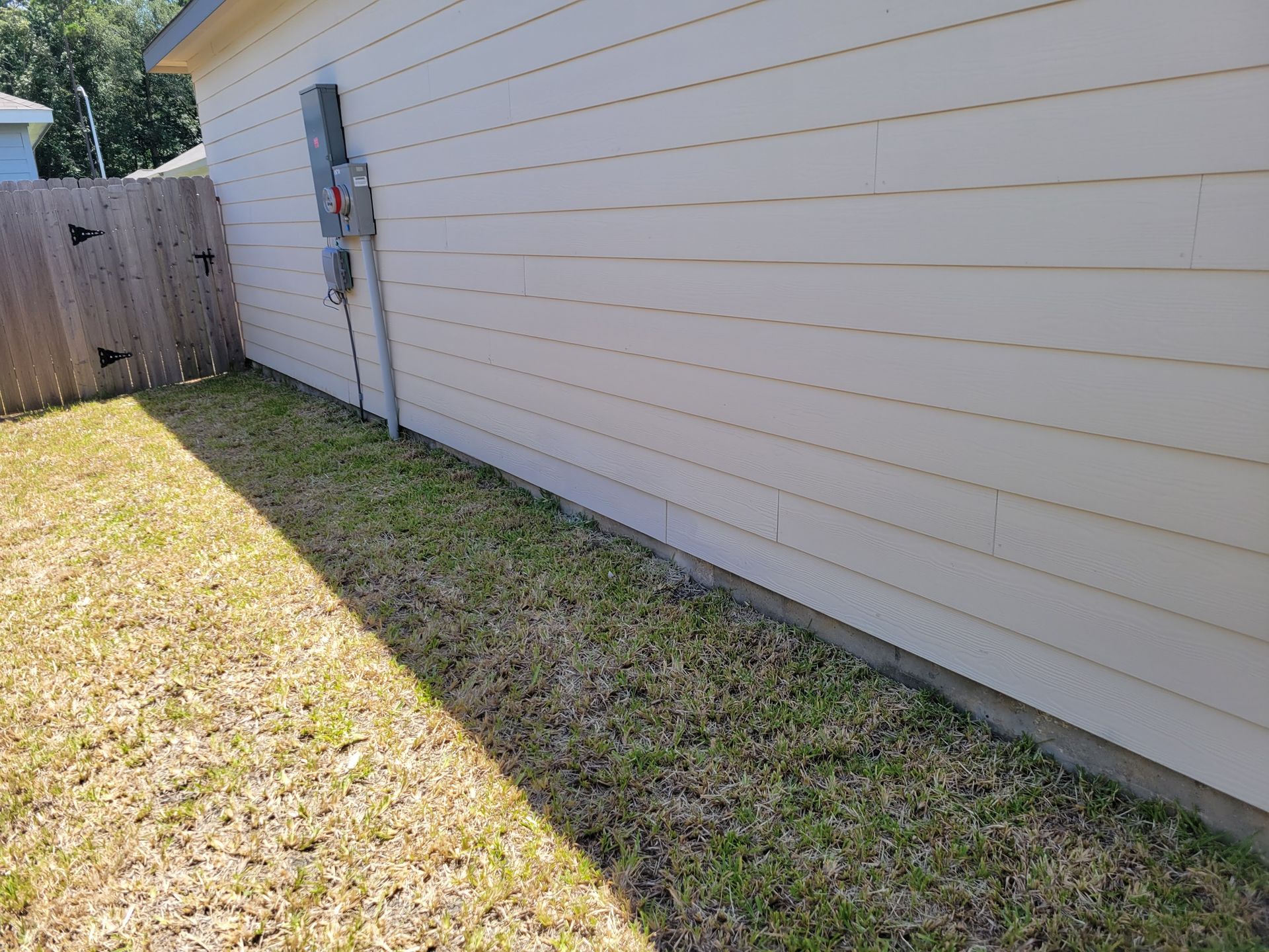 Grass strip next to a house with a wooden fence. Electrical box on the wall. Sunny day.