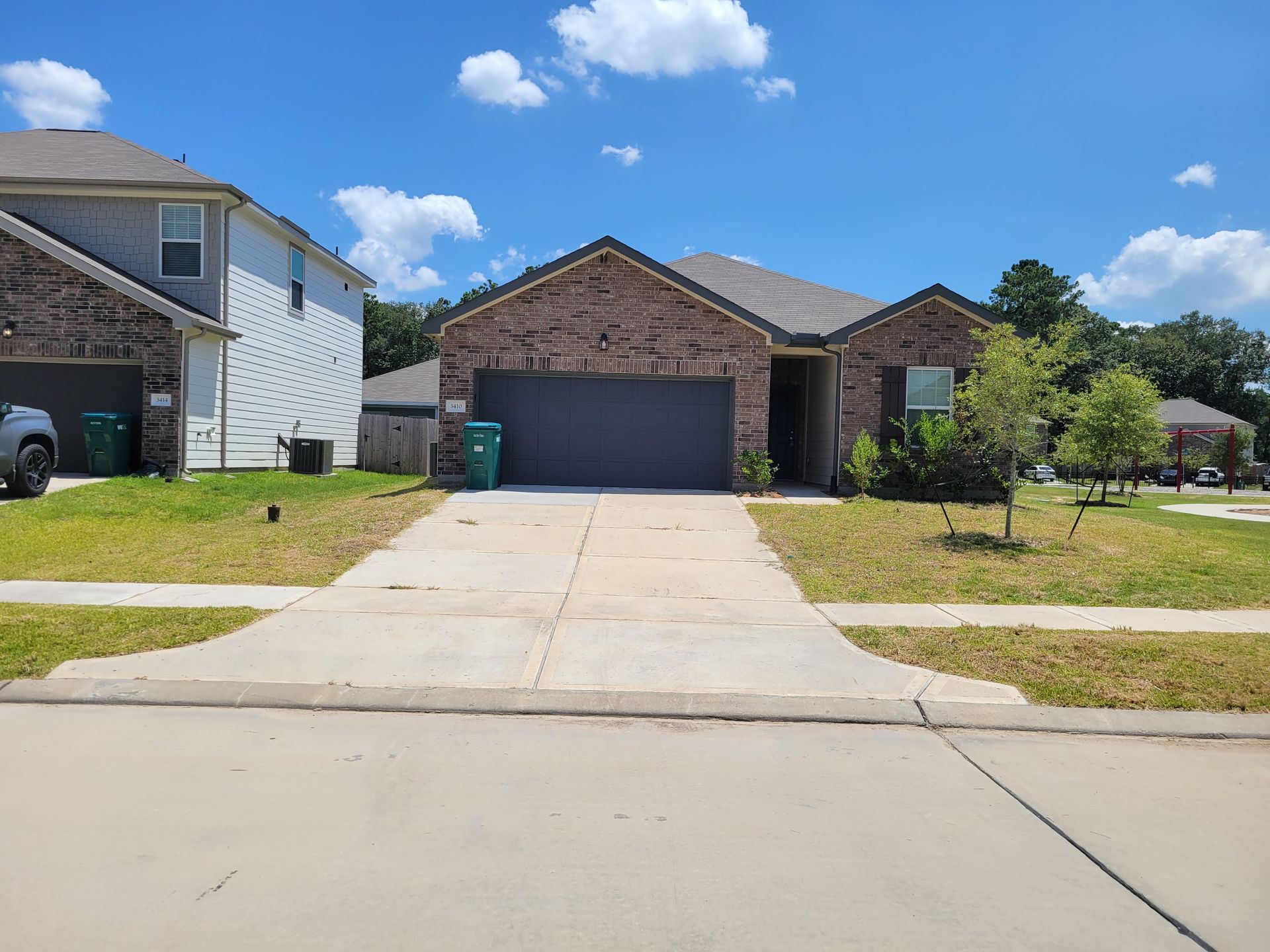 A brick home with a gray garage door sits on a concrete driveway, under a blue sky with fluffy clouds.