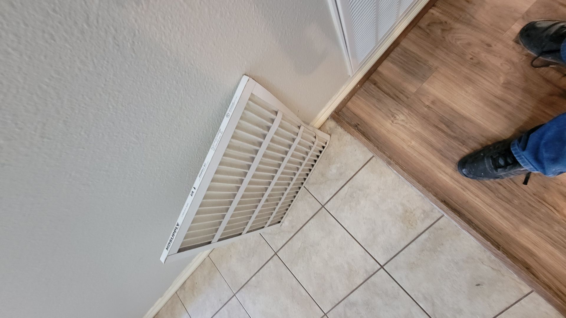 White air vent in a ceiling corner, adjacent to a wooden floor and tiled wall. A person's legs are visible.