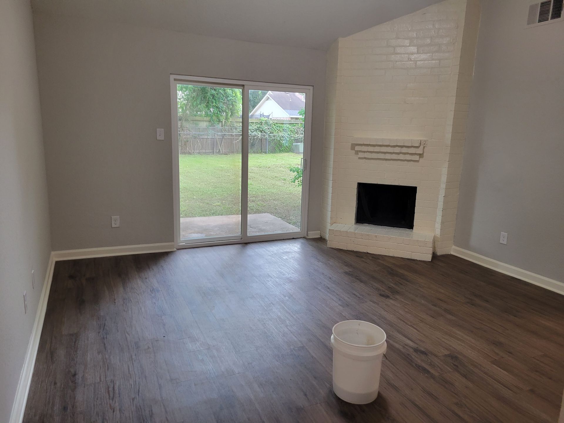 Empty living room with dark wood flooring, sliding glass door, white fireplace, and a bucket.