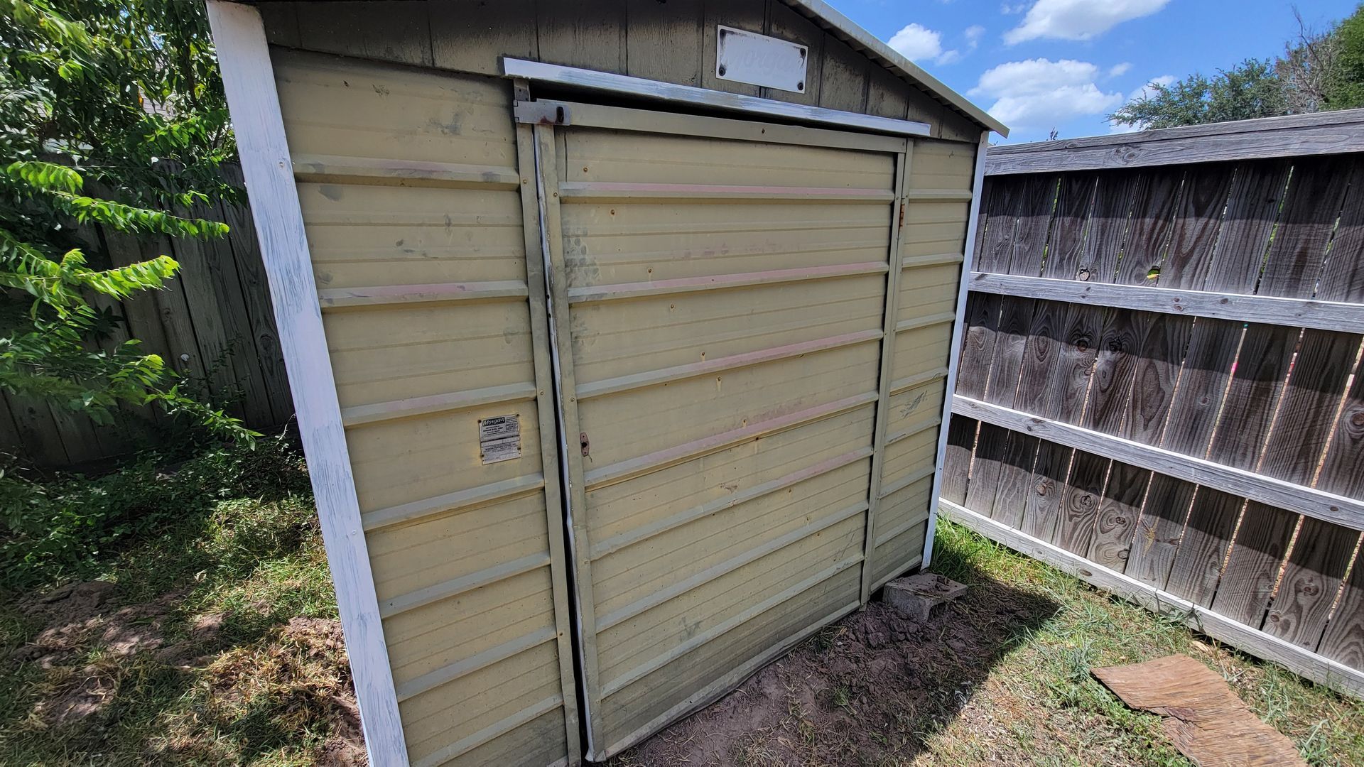A small, weathered shed with a sliding door in a grassy backyard, next to a wooden fence.