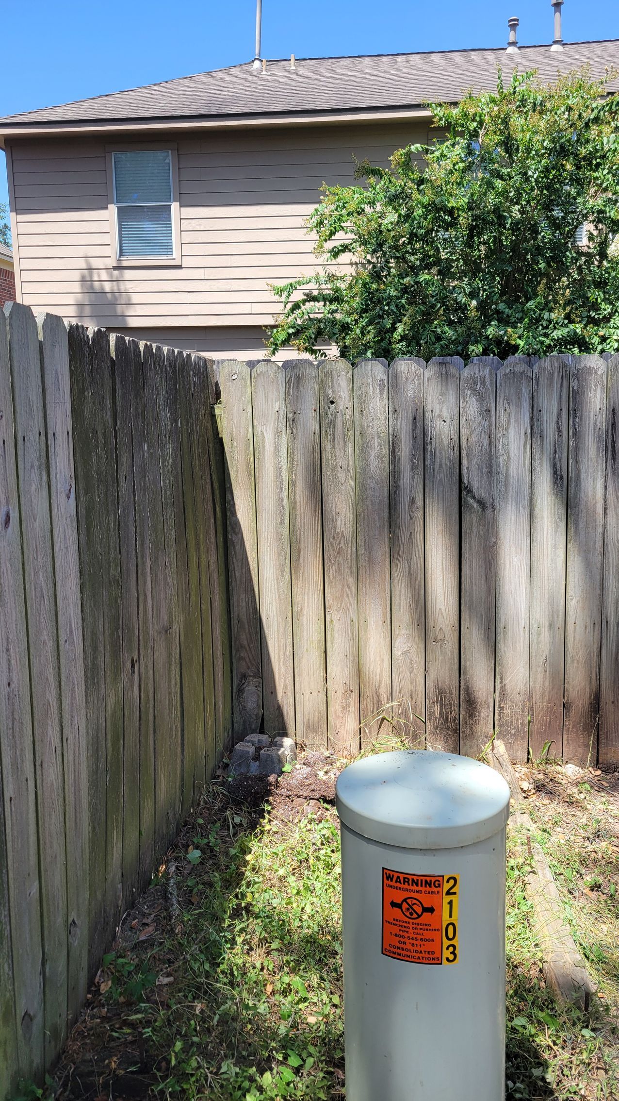 Wooden fence next to a white utility cylinder, with a two-story building in the background.