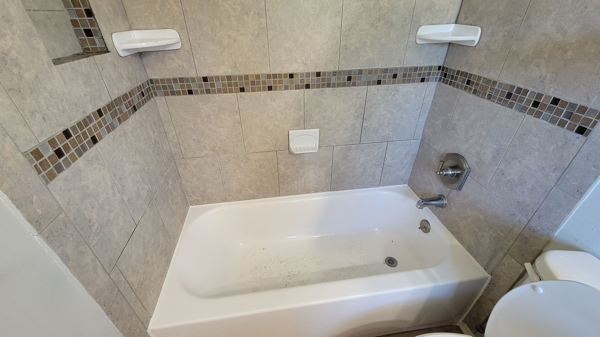 A small tiled bathroom with a white bathtub and toilet, two soap dishes, and a decorative tile border.