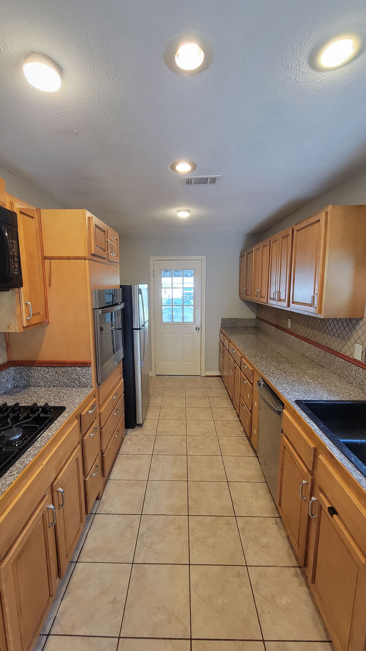 Long, narrow kitchen with light wood cabinets, granite countertops, and tile floor.