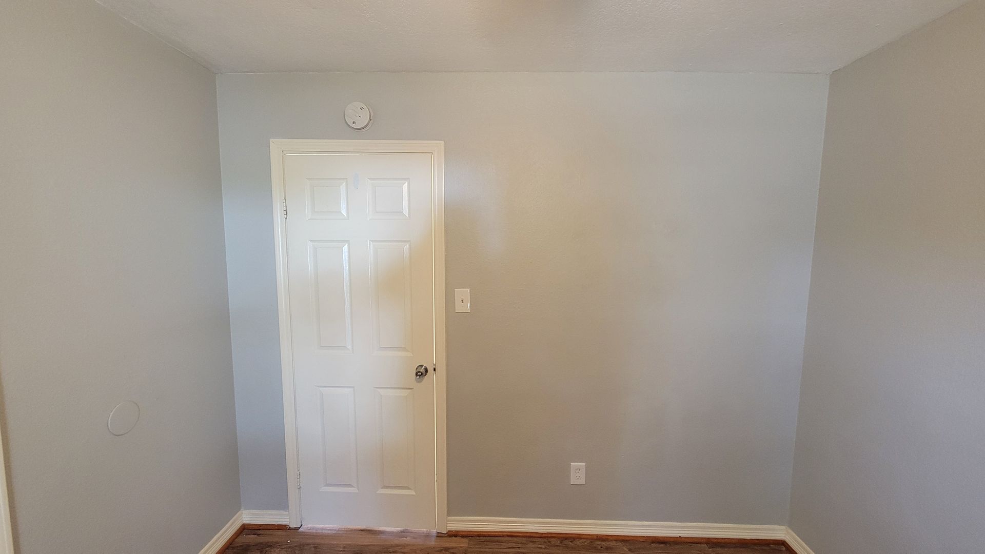 A light gray room with a white door centered on the wall and a dark carpet.