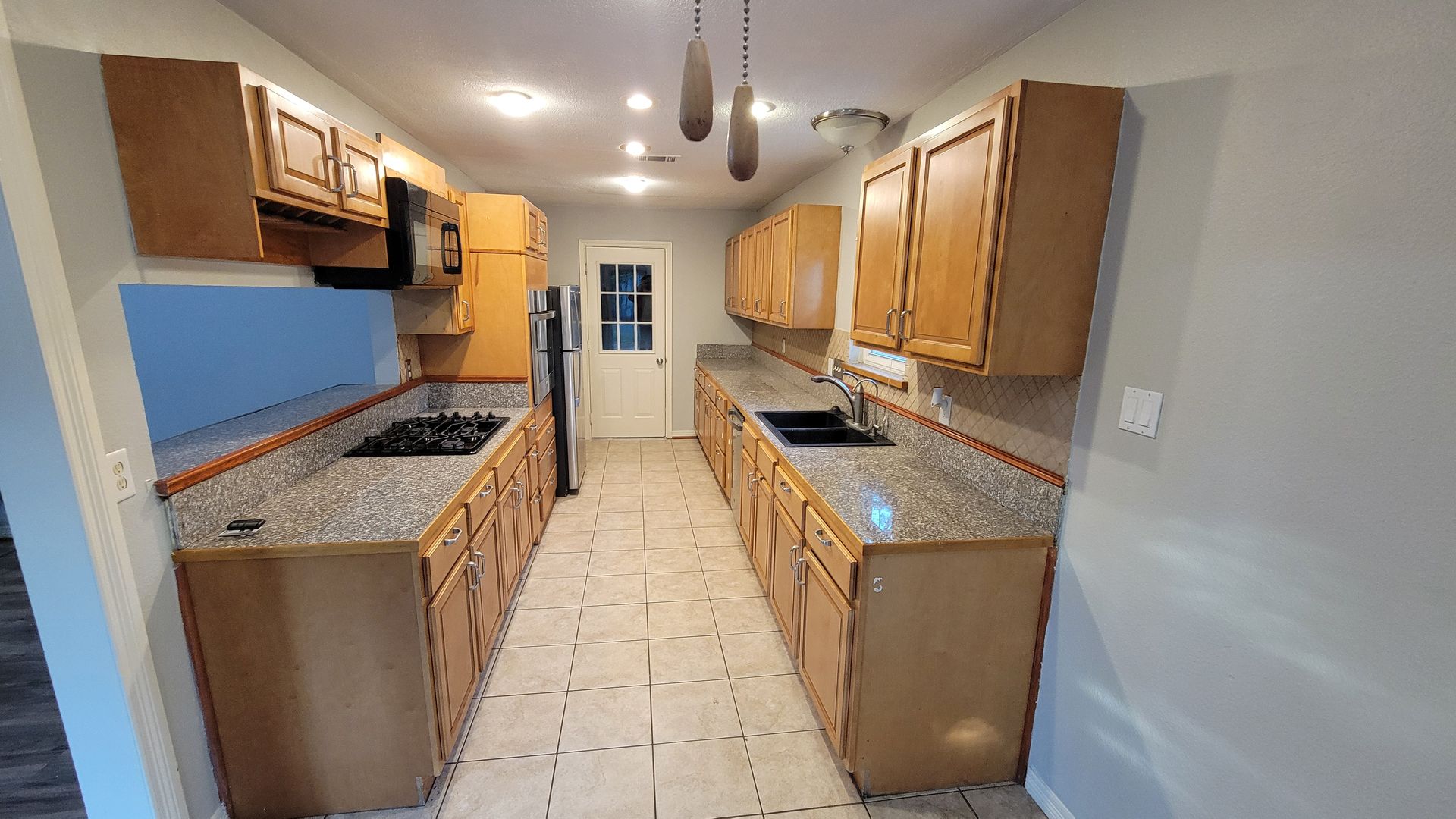 Kitchen with wooden cabinets, gray countertops, and tile flooring. A gas stovetop and sink are visible.